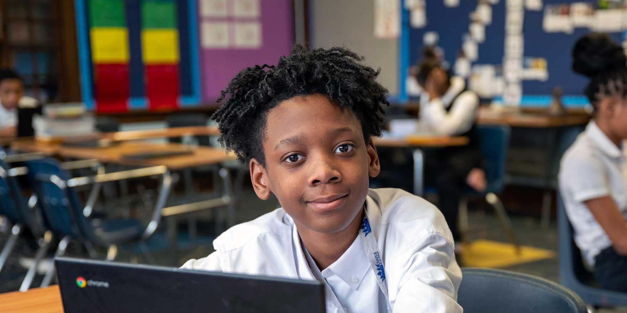 Student smiling at the camera from a classroom desk with a laptop.