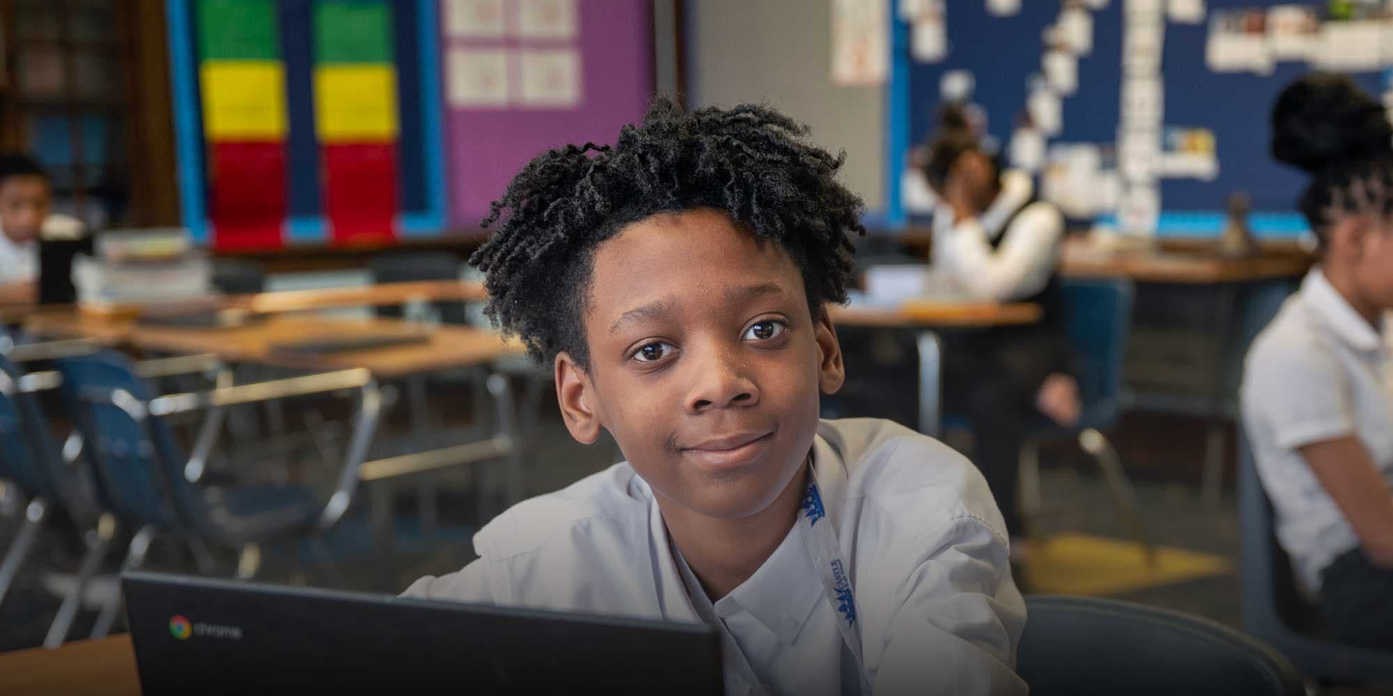 Student smiling at the camera while working on a laptop in a classroom.